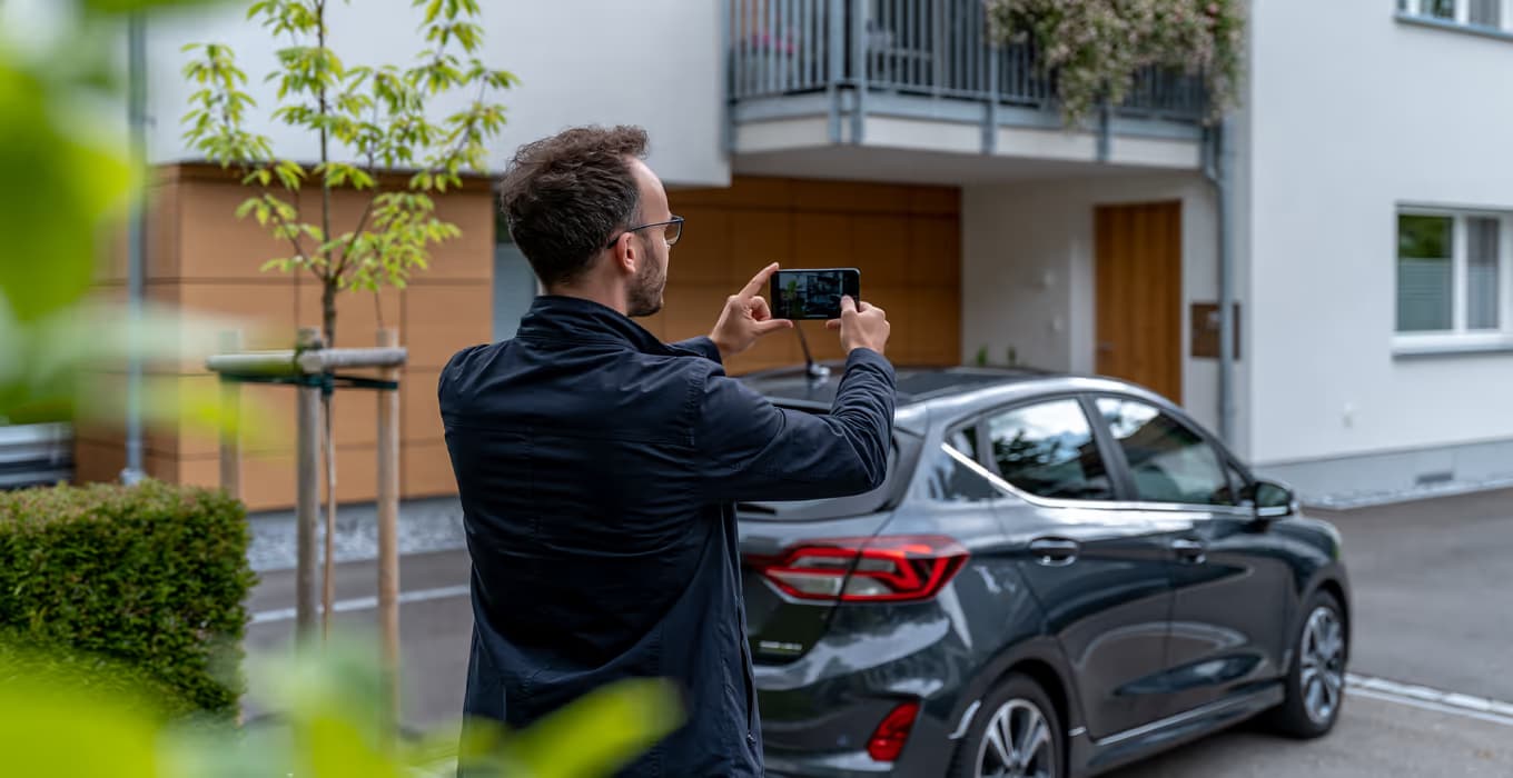 Mann fotografiert mit dem Smartphone ein dunkles geparktes Auto in einem Wohnhof